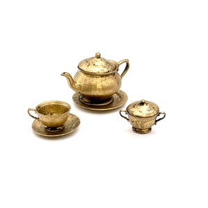 May include: A gold-toned miniature tea set with a teapot, cup and saucer, and a creamer. The set is made of metal and has a hammered finish.