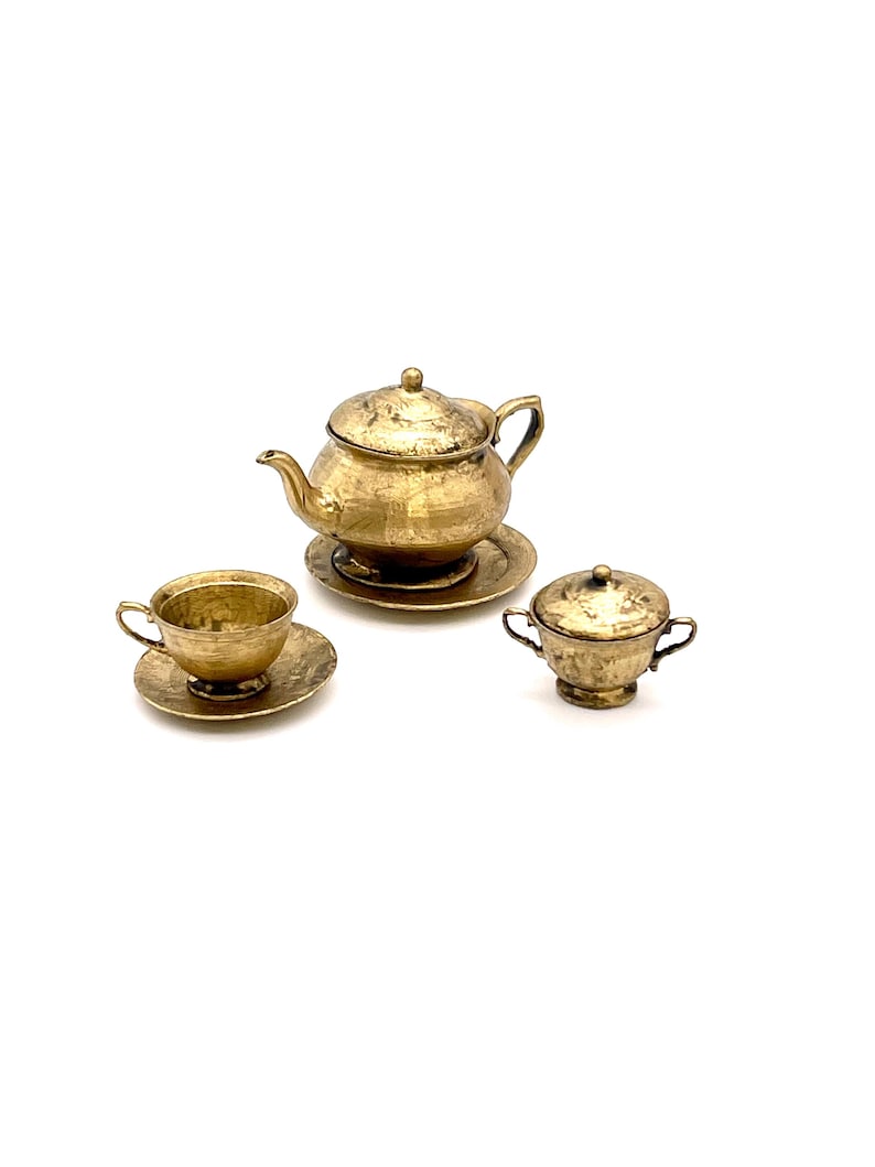 May include: A gold-toned miniature tea set with a teapot, cup and saucer, and a creamer. The set is made of metal and has a hammered finish.