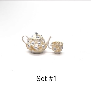 May include: A miniature teapot and matching teacup set featuring a textured cream-colored surface with raised, metallic silver heart accents. The teapot has a lid with a silver knob. The text Set #1 is printed in black below the items.