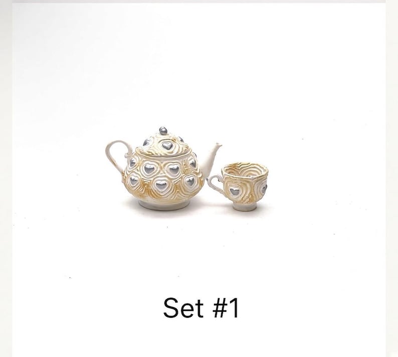 May include: A miniature teapot and matching teacup set featuring a textured cream-colored surface with raised, metallic silver heart accents. The teapot has a lid with a silver knob. The text Set #1 is printed in black below the items.