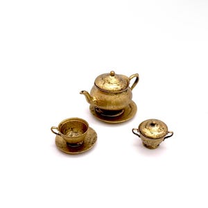 May include: A miniature gold tea set with a teapot, cup and saucer, and a creamer. The set is made of metal and has a hammered finish.