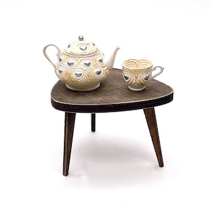 May include: A miniature ceramic teapot and matching teacup featuring a cream-colored base with swirling textures and raised silver heart accents. The set is displayed on a small, dark brown wooden three-legged table with a triangular top.