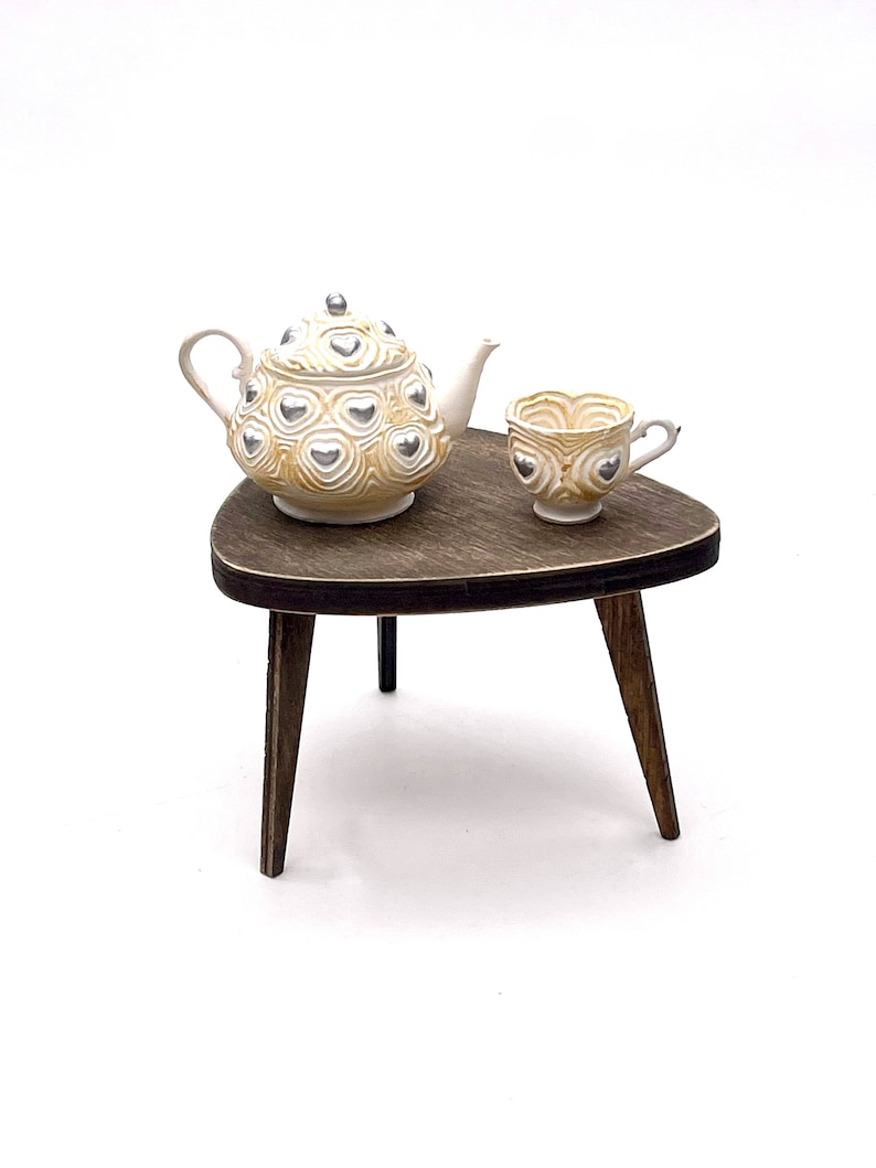 May include: A miniature ceramic teapot and matching teacup featuring a cream-colored base with swirling textures and raised silver heart accents. The set is displayed on a small, dark brown wooden three-legged table with a triangular top.