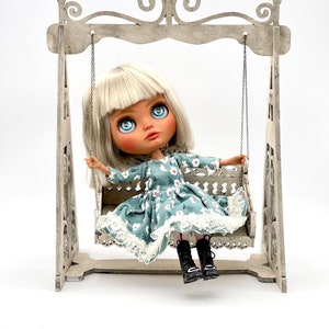 May include: A doll with blonde hair and blue eyes sits on a white wooden swing set. The doll is wearing a blue and white floral dress with a white lace trim and black boots.