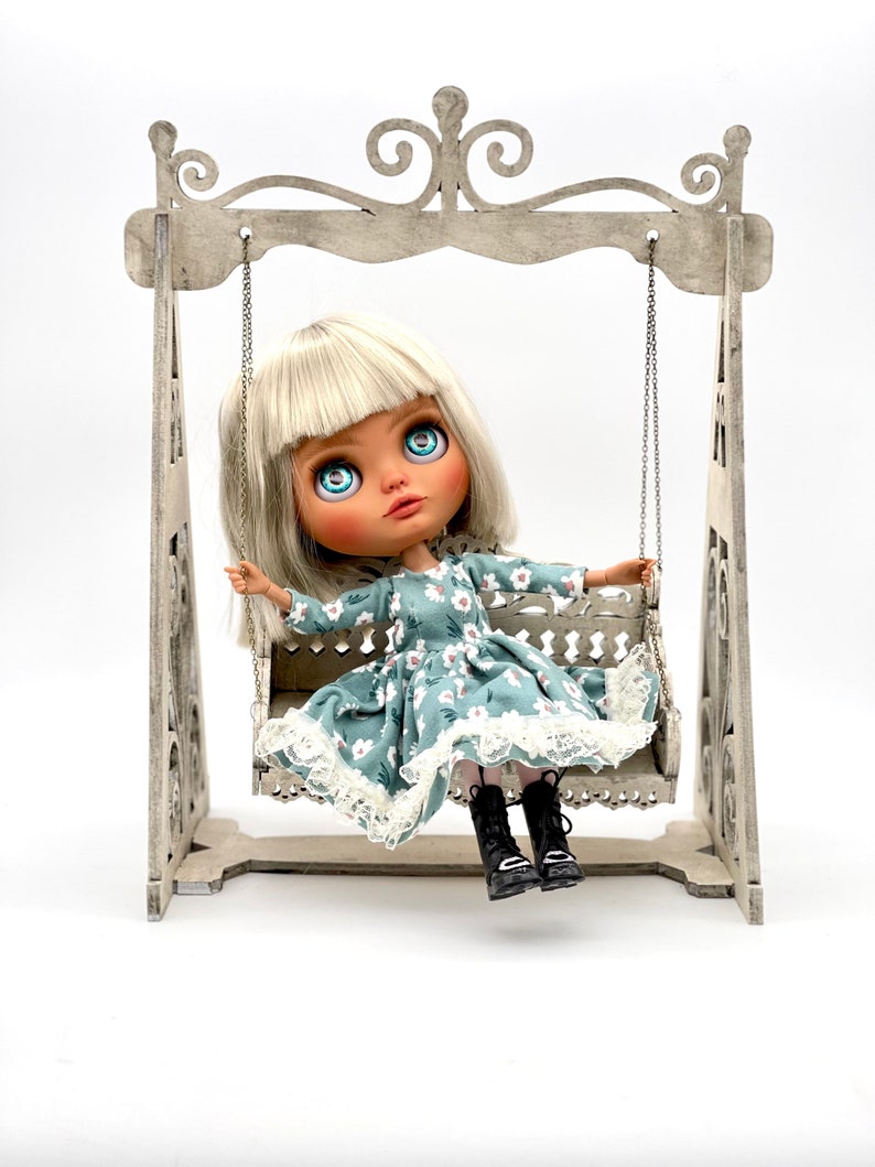 May include: A doll with blonde hair and blue eyes sits on a white wooden swing set. The doll is wearing a blue and white floral dress with a white lace trim and black boots.