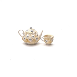 May include: A white teapot and teacup set with a gold and silver heart design.