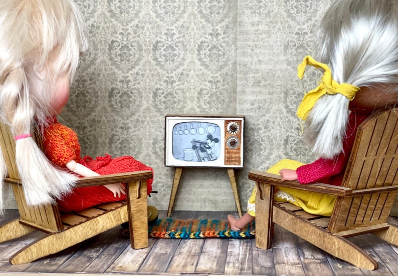 May include: Miniature scene with two dolls in wooden chairs watching a vintage TV. The TV shows a black and white cartoon. The dolls are wearing colorful outfits. The room has a patterned wallpaper and a small rug.