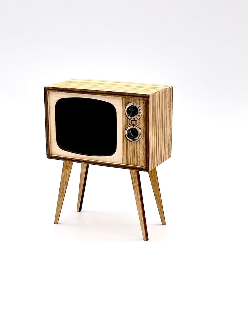 May include: A miniature wooden television with a retro design. The TV has a black screen, two circular knobs, and is supported by four angled legs. The wood has a light brown color with vertical graining.