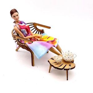 May include: A doll with brown hair wearing a sleeveless dress with a colorful abstract pattern of yellow, orange, purple, and blue. The doll sits in a curved wooden chair next to a small wooden table shaped like a leaf, holding a white teapot and cup.