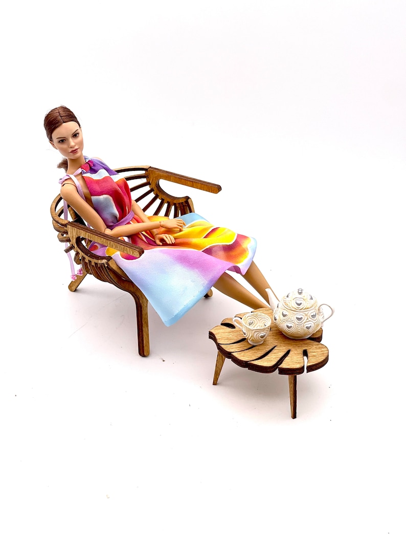 May include: A doll with brown hair wearing a sleeveless dress with a colorful abstract pattern of yellow, orange, purple, and blue. The doll sits in a curved wooden chair next to a small wooden table shaped like a leaf, holding a white teapot and cup.