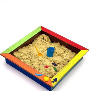 May include: A small wooden sandbox with a colorful rainbow border. The sandbox is filled with sand and includes a small blue bucket, a yellow shovel, and several small plastic fish toys.