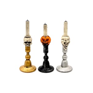 May include: Three Halloween themed candle holders. The candle holders are made of metal and have a skull, pumpkin, and skull design. The candle holders are painted gold, black, and silver.