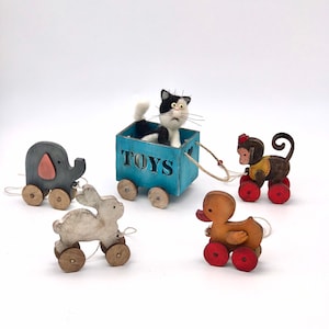 May include: A set of five wooden toy animals on wheels. The animals include a cat, a duck, a rabbit, an elephant, and a monkey. The cat is in a blue wagon with the word "TOYS" written on the side. The toys are all painted in bright colours.