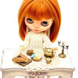 May include: A doll with red hair sits at a white table set with a crocheted tablecloth. The table is decorated with a teapot, teacups, pastries, and a candlestick.