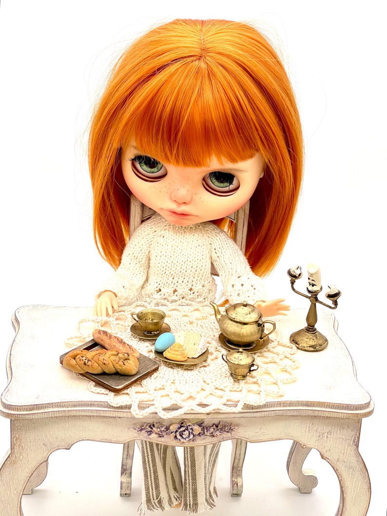 May include: A doll with red hair sits at a white table set with a crocheted tablecloth. The table is decorated with a teapot, teacups, pastries, and a candlestick.