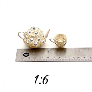 May include: A miniature teapot and teacup set featuring a cream-colored base with a raised, swirling texture and silver heart accents. The items are positioned next to a metal ruler showing a scale of 1:6, with the teapot measuring about 1.5 inches.