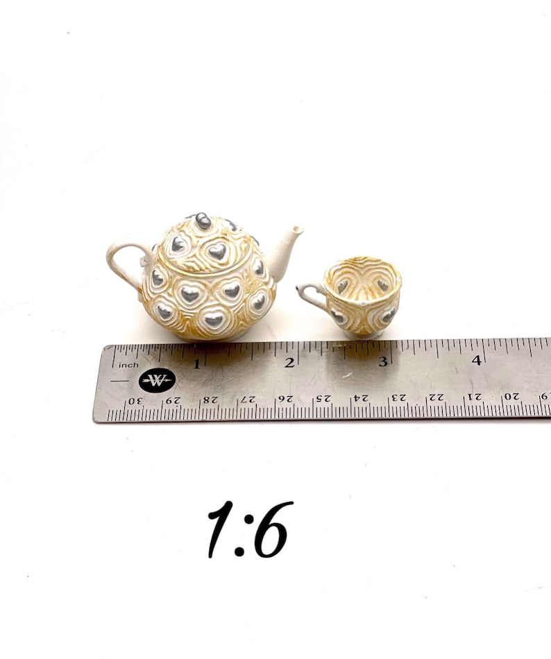 May include: A miniature teapot and teacup set featuring a cream-colored base with a raised, swirling texture and silver heart accents. The items are positioned next to a metal ruler showing a scale of 1:6, with the teapot measuring about 1.5 inches.