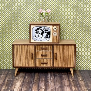 May include: A miniature wooden television set and matching cabinet. The TV displays a black and white cartoon. The cabinet has three drawers and two doors with vertical wood grain. A small vase of pink flowers sits atop the TV.