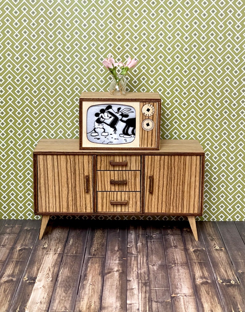 May include: A miniature wooden television set and matching cabinet. The TV displays a black and white cartoon. The cabinet has three drawers and two doors with vertical wood grain. A small vase of pink flowers sits atop the TV.