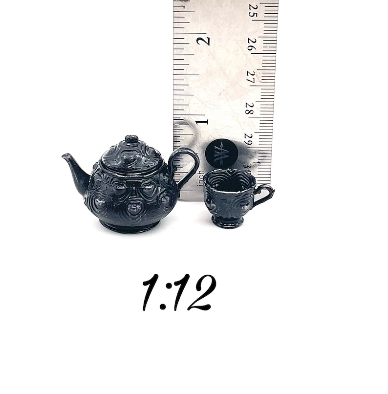 May include: A miniature black teapot and matching teacup featuring a raised heart pattern, displayed next to a ruler showing a height of approximately 1 inch for the teapot. The text 1:12 is printed below the items on a white background.