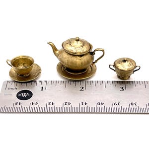 May include: A miniature gold-colored tea set with a teapot, creamer, and cup and saucer. The set is about 2 inches tall and is sitting on a ruler for scale.
