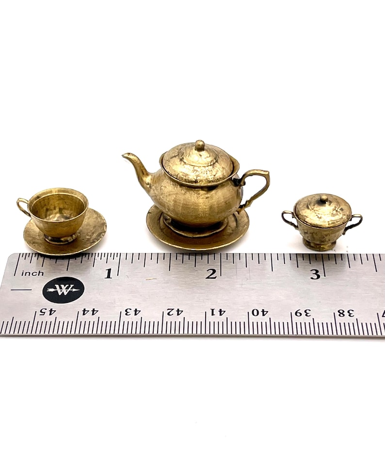 May include: A miniature gold-colored tea set with a teapot, creamer, and cup and saucer. The set is about 2 inches tall and is sitting on a ruler for scale.