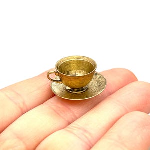 May include: A miniature gold teacup and saucer set. The cup and saucer are both made of metal and have a slightly textured surface.