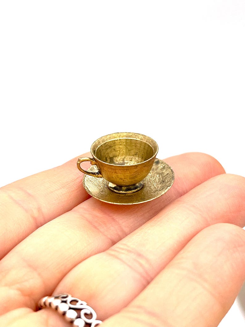 May include: A miniature gold teacup and saucer set. The cup and saucer are both made of metal and have a slightly textured surface.