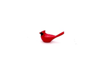 May include: A red cardinal figurine with a black beak and black eyes. The figurine is perched on a white surface.