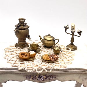 May include: A miniature tea set with a samovar, teapot, creamer, and two cups and saucers. The set is arranged on a white crocheted doily on a white table with a floral design.