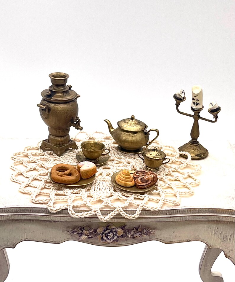 May include: A miniature tea set with a samovar, teapot, creamer, and two cups and saucers. The set is arranged on a white crocheted doily on a white table with a floral design.