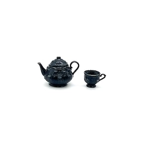 May include: A miniature black teapot and a matching teacup set. Both items feature a raised, embossed pattern across their surfaces. The teapot has a curved spout, a lid with a small knob, and a handle. The teacup has a small handle and a pedestal base.
