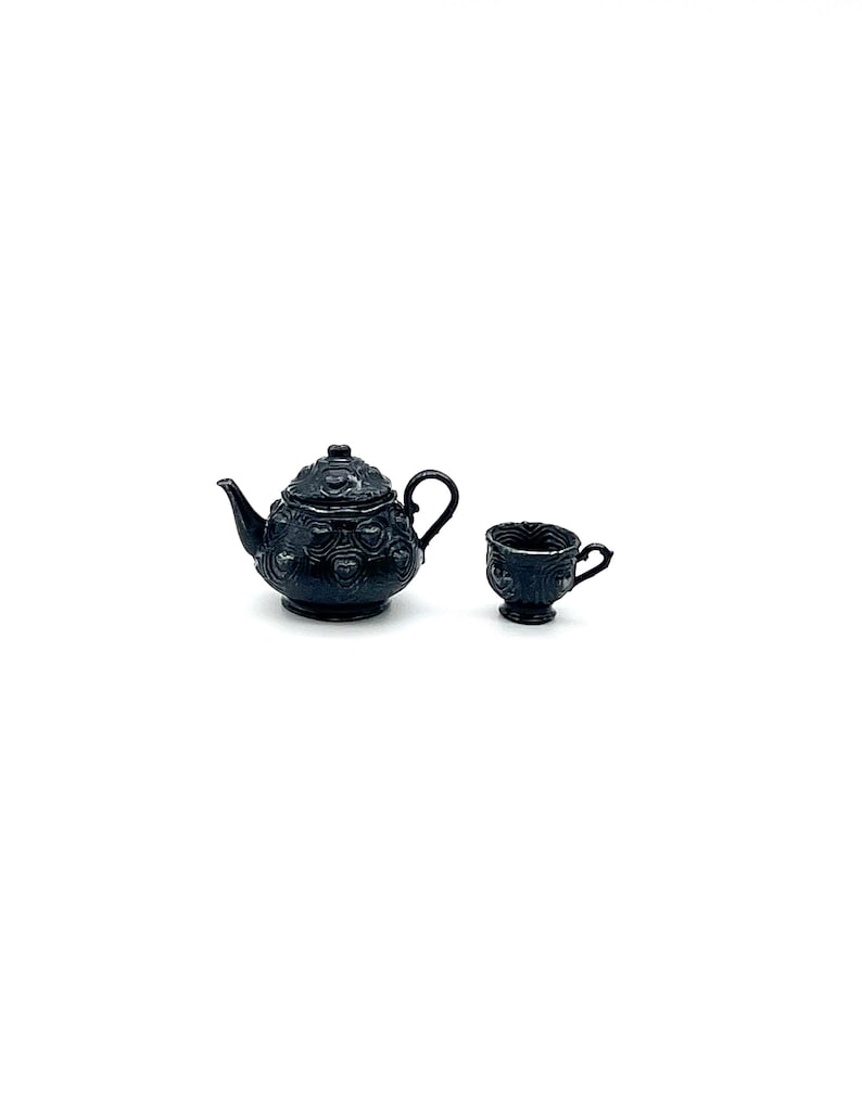 May include: A miniature black teapot and a matching teacup set. Both items feature a raised, embossed pattern across their surfaces. The teapot has a curved spout, a lid with a small knob, and a handle. The teacup has a small handle and a pedestal base.