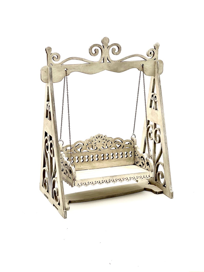 May include: A white wooden swing set with intricate carvings and a decorative top. The swing is suspended from chains and has a white wooden seat.