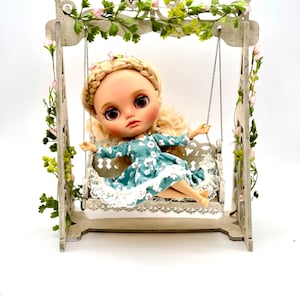 May include: A doll with blonde hair and a blue floral dress sits on a white swing set with green vines. The swing set is decorated with pink flowers.