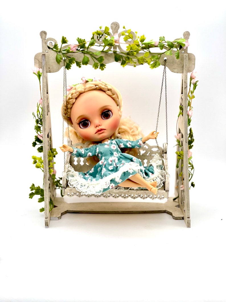 May include: A doll with blonde hair and a blue floral dress sits on a white swing set with green vines. The swing set is decorated with pink flowers.