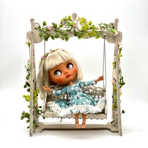 May include: A doll wearing a blue and white floral dress sits on a white wooden swing set with a green vine and pink flowers hanging from the top.