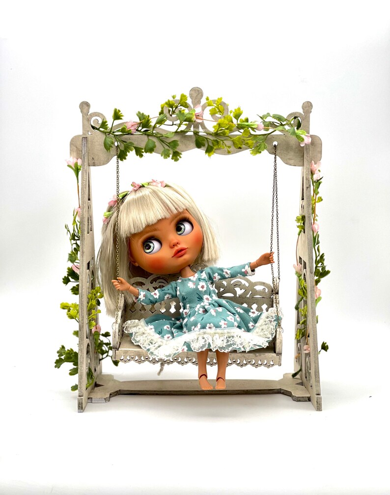 May include: A doll wearing a blue and white floral dress sits on a white wooden swing set with a green vine and pink flowers hanging from the top.