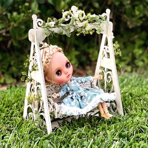 May include: A doll with blonde hair and blue eyes sits on a white swing set with a floral pattern dress. The swing set is decorated with greenery and white flowers.