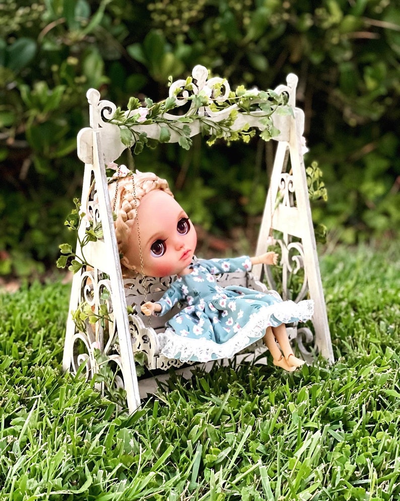 May include: A doll with blonde hair and blue eyes sits on a white swing set with a floral pattern dress. The swing set is decorated with greenery and white flowers.