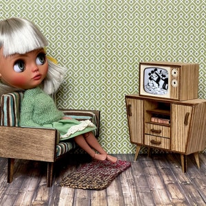 May include: A doll sits in a miniature mid-century modern chair, wearing a green sweater and dress. A miniature wooden TV and cabinet set displays a black and white cartoon. A small rug is on the floor.