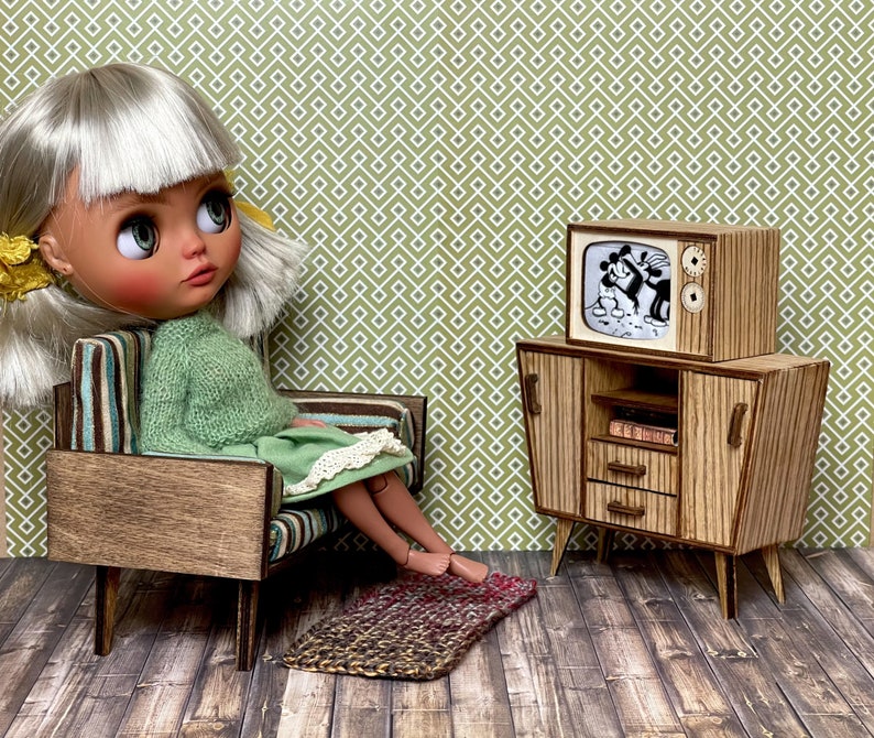 May include: A doll sits in a miniature mid-century modern chair, wearing a green sweater and dress. A miniature wooden TV and cabinet set displays a black and white cartoon. A small rug is on the floor.