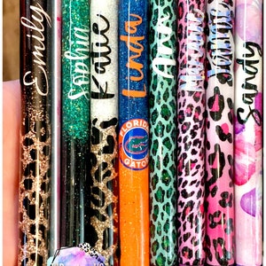 May include: Nine personalized pens with glitter and various patterns, including leopard print, glitter, and a Florida Gators logo. The pens are labeled with names: Emily, Sophia, Katie, Linda, Anna, Madison, Jasmine, and Sandy.