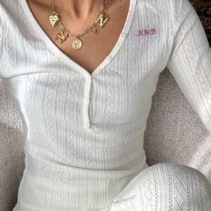 Puede incluir: Conjunto de pijama blanco de manga larga con un delicado diseño calado. La parte superior presenta un cuello en V con tres botones y las iniciales "NMB" bordadas en rosa. Un collar dorado con colgantes añade un toque de elegancia.