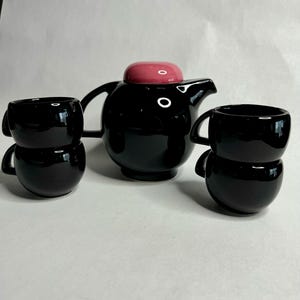 Op de afbeelding: Een zwarte theepot met een roze deksel en twee zwarte theekopjes die op elkaar gestapeld zijn. De theepot en de kopjes zijn gemaakt van keramiek.