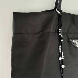 以下が含まれることがあります： 黒のナイロン製のトートバッグで、黒と白のビーズのストラップが付いています。ストラップには白いビーズと、白い正方形が付いた黒いビーズが付いています。