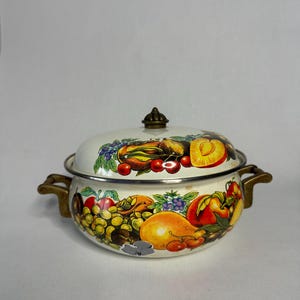 Puede incluir: Olla de esmalte blanco con tapa que presenta un diseño de frutas coloridas. La olla tiene dos asas de latón y una perilla de latón en la tapa.