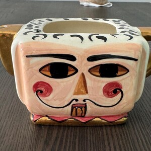 Op de afbeelding: Een keramische theepot met een fantasievol gezicht erop geschilderd. De theepot is wit met zwarte en gouden accenten. Het gezicht heeft bruine ogen, een bruine neus en een roze mond met een snor. De theepot heeft een gouden handvat en tuit.