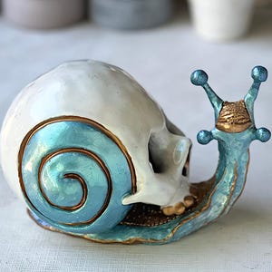 May include: A ceramic snail sculpture with a skull-shaped shell. The shell is white with a blue spiral design and gold trim. The snail's body is blue with gold accents, and it has blue antennae.