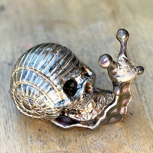 Op de afbeelding: Een decoratief slakkenbeeld met een schedelvormige schelp en een gedetailleerd, getextureerd lichaam. De schelp is donkergrijs met gouden accenten, en het lichaam is van metaalgoud.